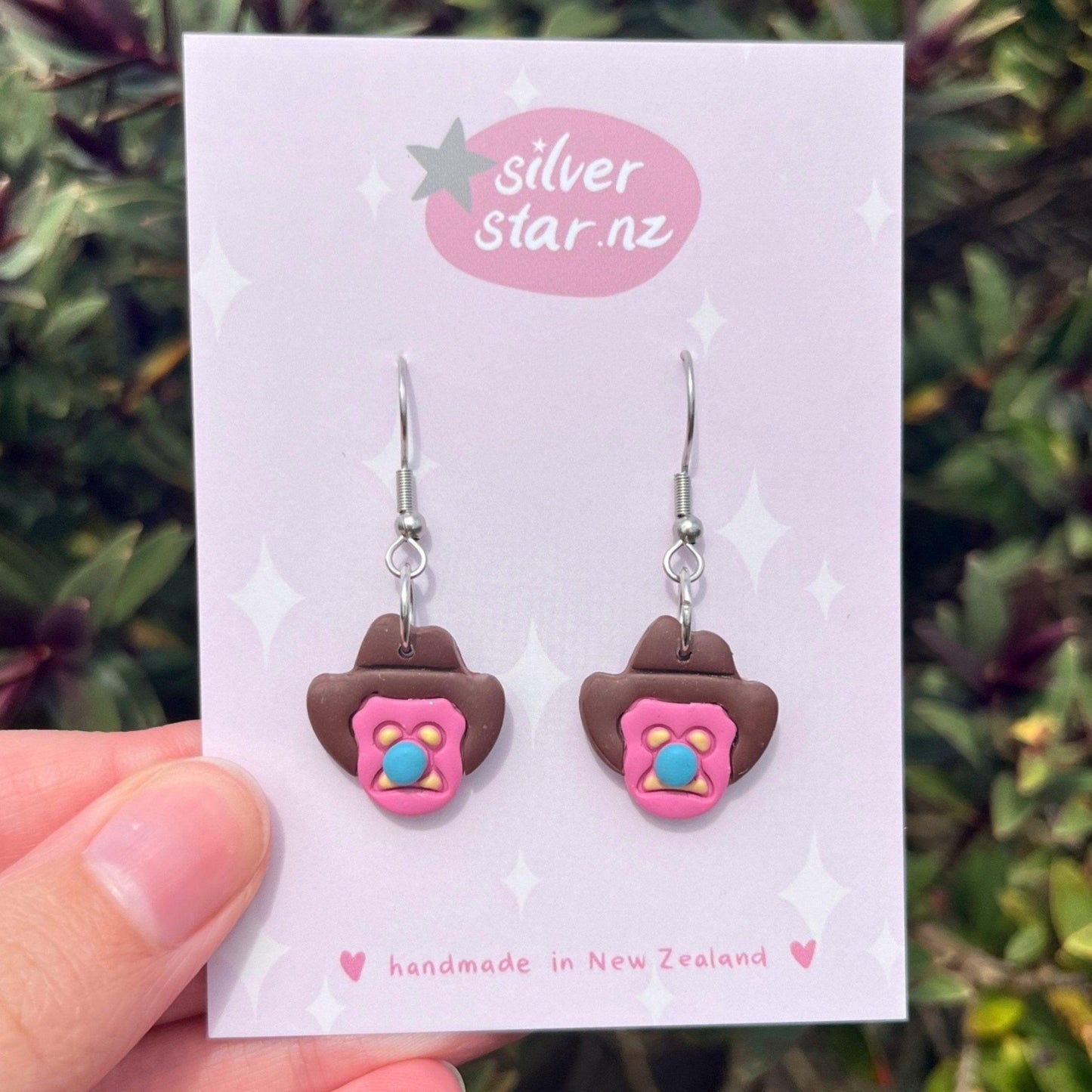 A hand holds a card with Bubble 'O Will Polymer Clay Earrings by silverstar.nz. These handmade earrings feature cowboy hat shapes with pink and blue accents and stainless steel hooks, crafted in New Zealand.