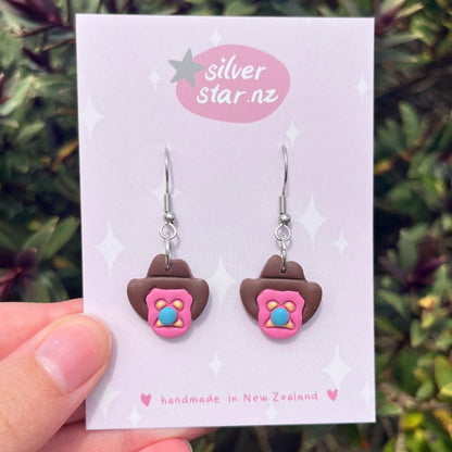 A hand holds a card with Bubble 'O Will Polymer Clay Earrings by silverstar.nz. These handmade earrings feature cowboy hat shapes with pink and blue accents and stainless steel hooks, crafted in New Zealand.