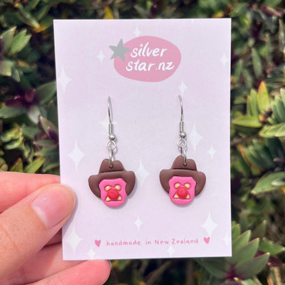 A hand holds a display card from silverstar.nz showing Bubble 'O Will Polymer Clay Earrings shaped like pink frosted biscuits with yellow centers and cowboy hats, handmade in New Zealand on stainless steel hooks.