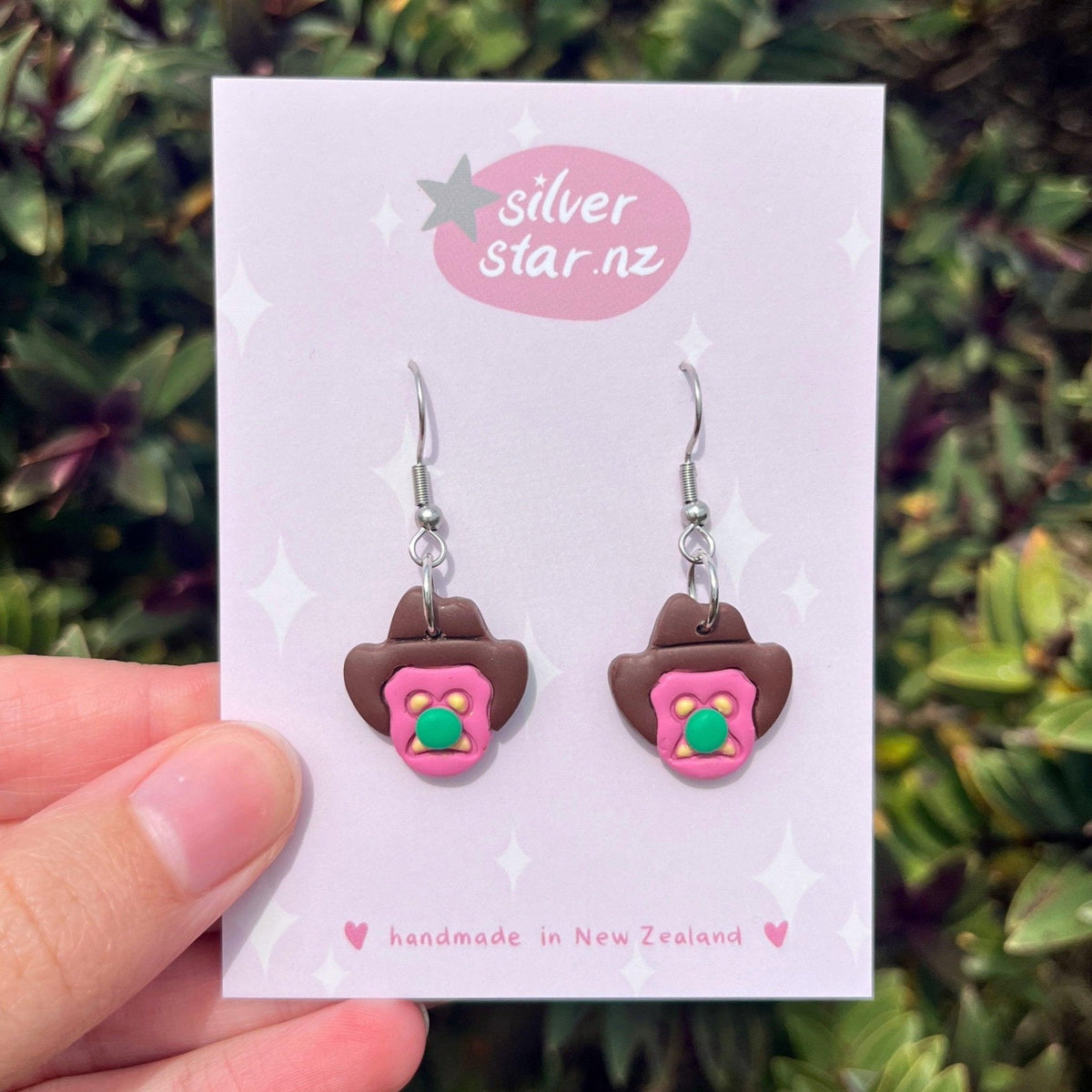A hand holds a card displaying Bubble 'O Will Polymer Clay Earrings by silverstar.nz. The handmade earrings are shaped like brown cowboy hats with pink and green floral centers. Green foliage appears blurred in the background.