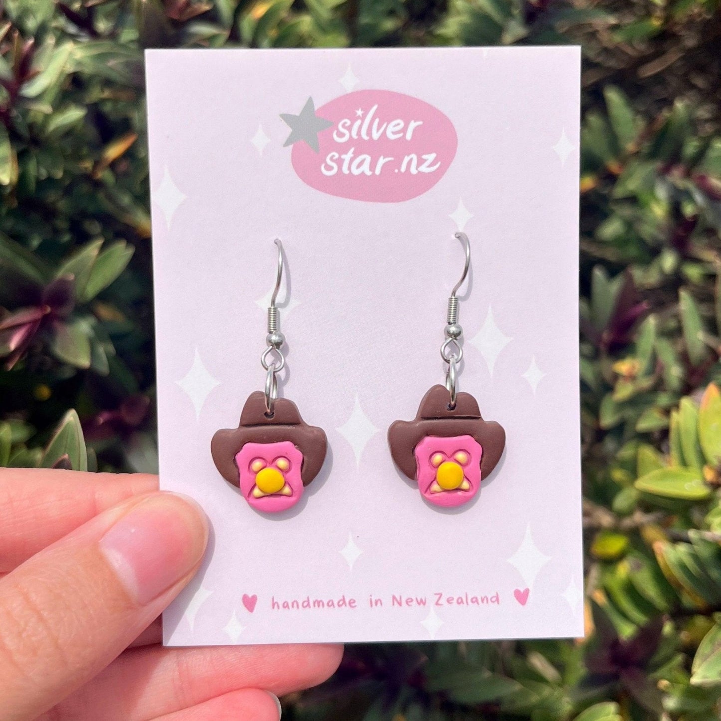 A hand holds a card with Bubble 'O Will Polymer Clay Earrings by silverstar.nz, featuring pink and brown cowboy hats adorned with yellow and pink flowers. The card says “silverstar.nz” and “handmade in New Zealand,” against green foliage.
