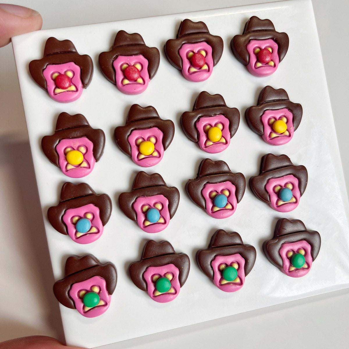 A white tray displays 16 Bubble 'O Will Polymer Clay Earrings by silverstar.nz—brown cowboy hat shapes with pink trim and a colorful candy center (yellow, blue, green, or pink), resembling playful handmade sweets.