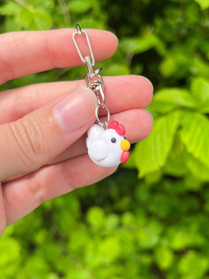 A hand displays the Stardew Chicken Polymer Clay Keyring by silverstar.nz, featuring a white chicken-shaped charm with a red comb and yellow beak—an ideal accessory for handmade keyring enthusiasts.