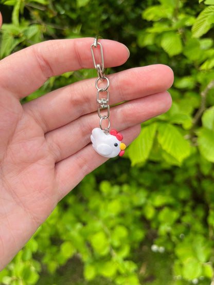 A hand holds the Stardew Chicken Polymer Clay Keyring by silverstar.nz, featuring a small white chicken charm with a red comb and yellow beak. Blurred green leaves in the background highlight this charming handmade accessory.