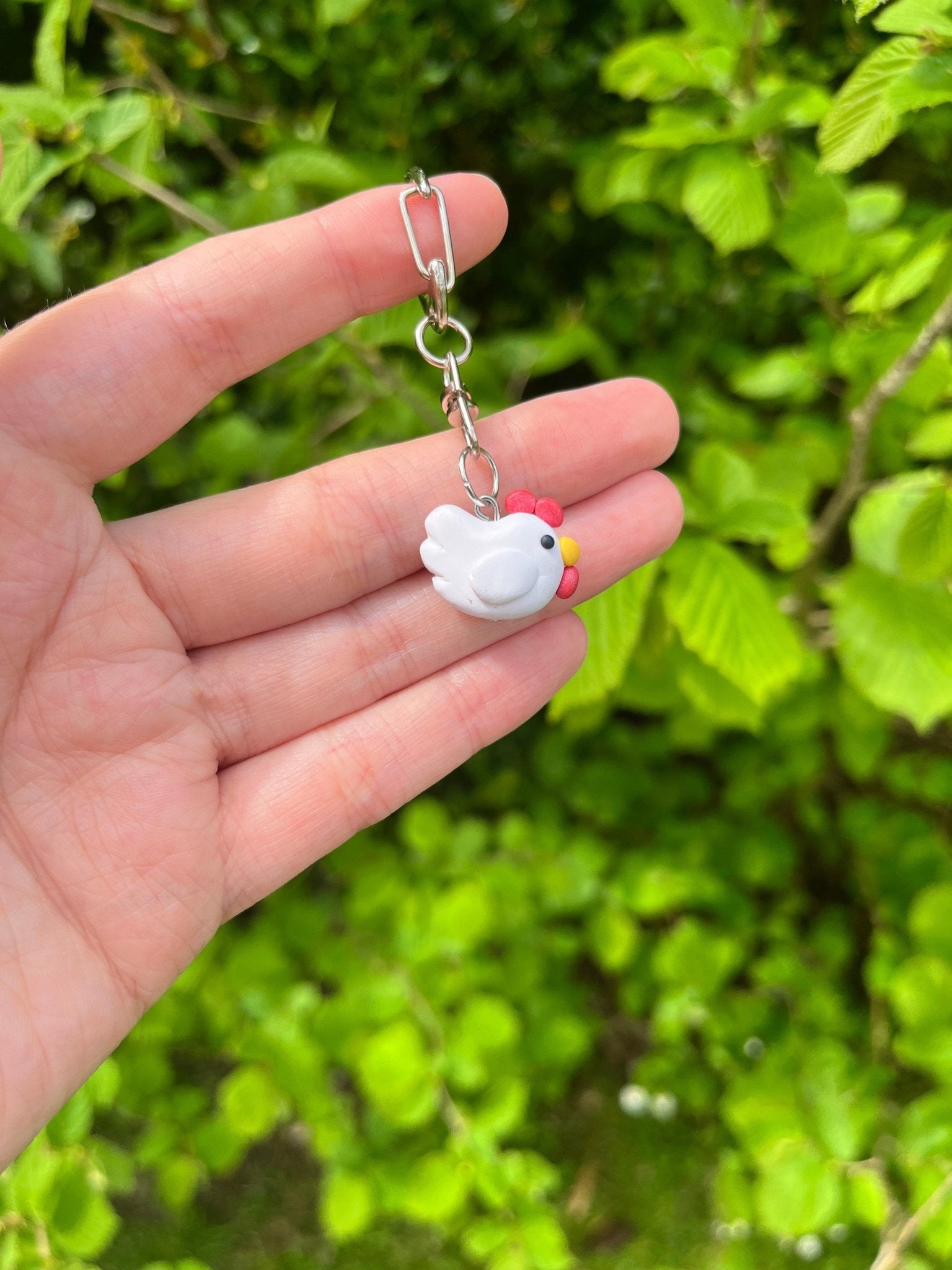 A hand holds the Stardew Chicken Polymer Clay Keyring by silverstar.nz, featuring a small white bird charm with a red beak and yellow eyes, set against green leafy plants.