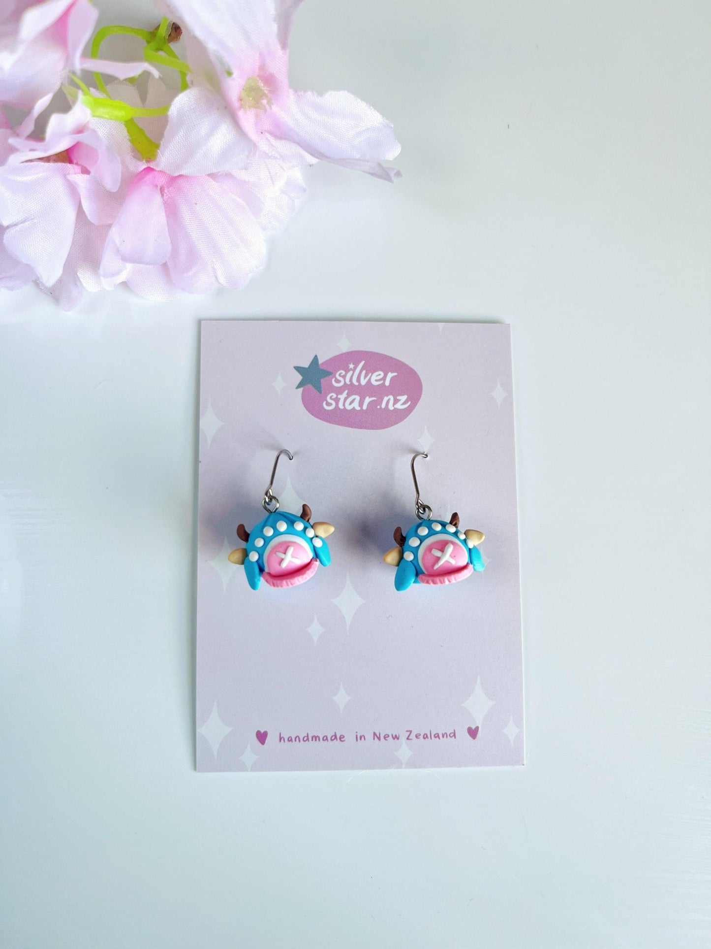 The Chopper Hat Polymer Clay Earrings by silverstar.nz are handmade blue and pink earrings with star and paw designs, shown on a branded card beside pink artificial flowers, and feature durable stainless steel hooks.