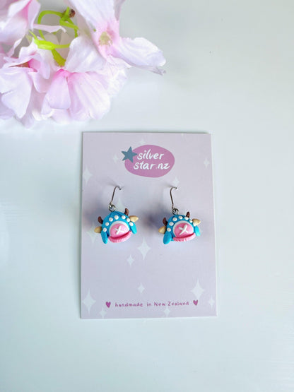 The Chopper Hat Polymer Clay Earrings by silverstar.nz are handmade blue and pink earrings with star and paw designs, shown on a branded card beside pink artificial flowers, and feature durable stainless steel hooks.