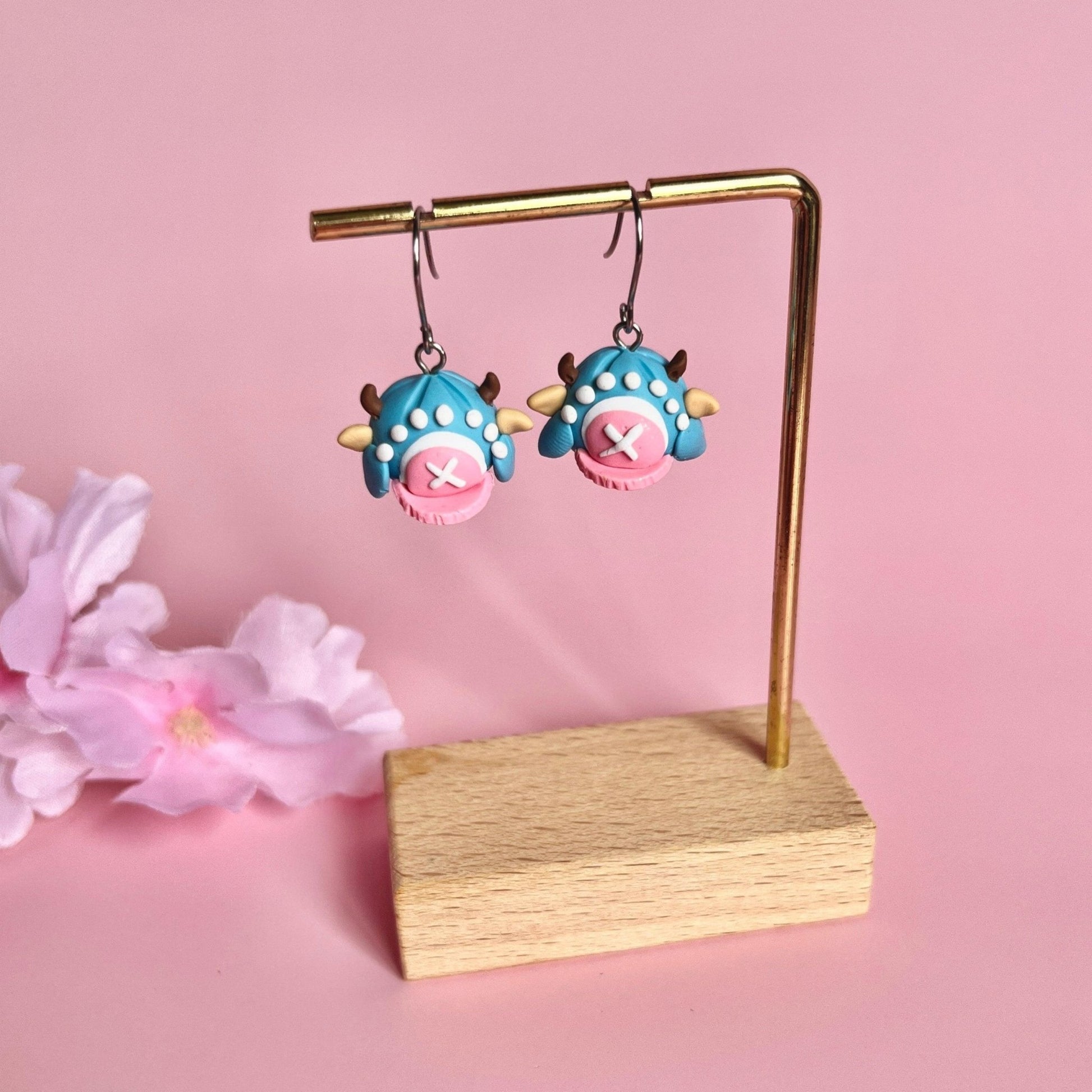 Chopper Hat Polymer Clay Earrings by silverstar.nz—whimsical handmade earrings with blue and pink monster designs, X-shaped mouths, horns, and stainless steel hooks. Displayed on a gold and wooden stand with pink flowers in the background.