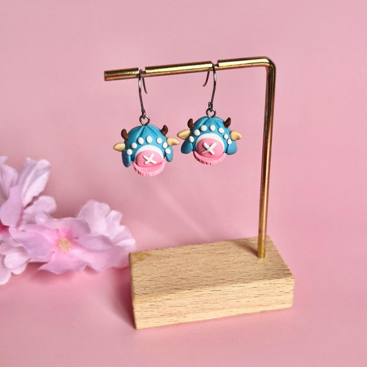 Chopper Hat Polymer Clay Earrings by silverstar.nz—whimsical handmade earrings with blue and pink monster designs, X-shaped mouths, horns, and stainless steel hooks. Displayed on a gold and wooden stand with pink flowers in the background.
