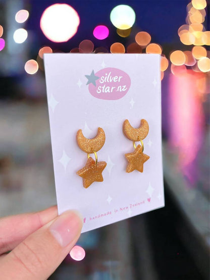 A hand presents Star + Moon Polymer Clay Earrings by silverstar.nz on a branded card. The design, featuring glittery gold crescent moons and stars, stands out against a blurred nightscape with colorful bokeh lights reflecting on water, highlighting the earrings whimsical charm.