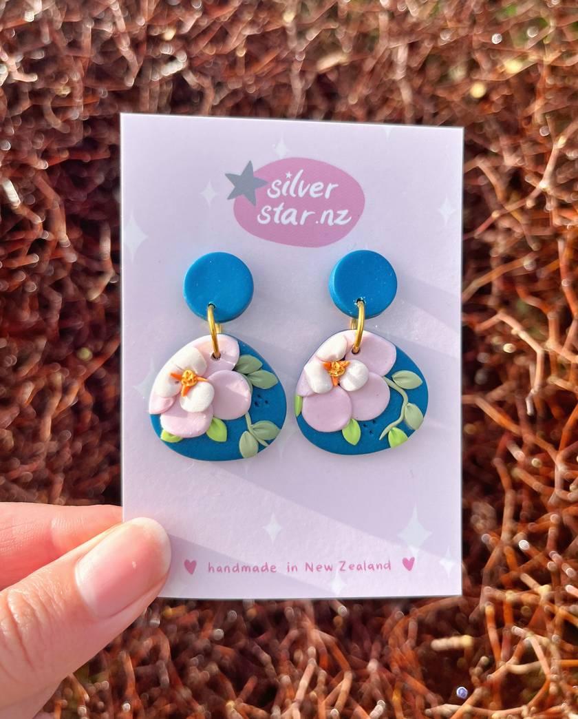 A hand holds a card with silverstar.nzs Blue Floral Polymer Clay Earrings, featuring blue flower shapes adorned with pink flowers and green leaves on stainless steel hooks, capturing New Zealands essence against a blurred natural foliage backdrop.