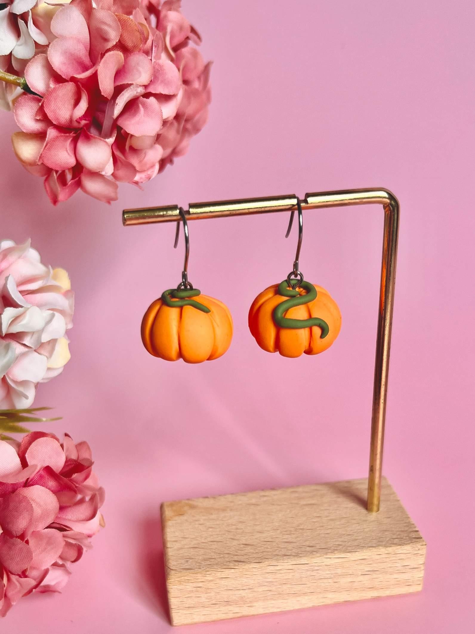 The Pumpkin Polymer Clay Earrings, featuring a distinctive mini pumpkin design, are displayed on a gold stand with a wooden base. These handmade earrings are set against a pink backdrop embellished with clusters of pink flowers on either side.